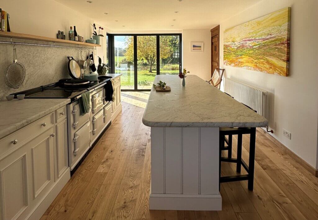 Bright modern kitchen with oak hardwood flooring, white marble worktops, and large glass doors opening to a sunny garden.